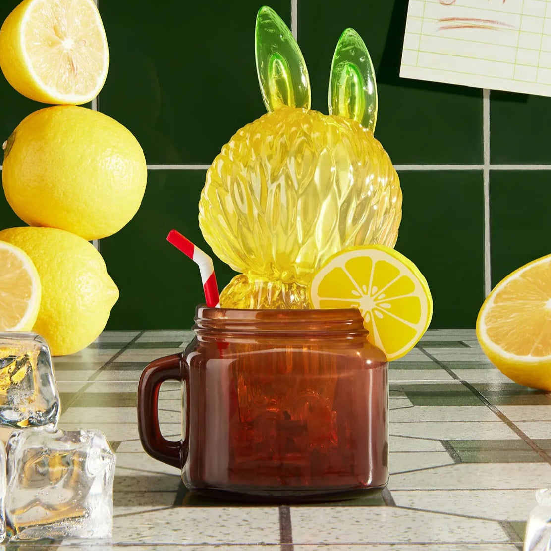Lemon-shaped drink mixer with rabbit ears in a brown mug, surrounded by lemons on a tiled countertop.